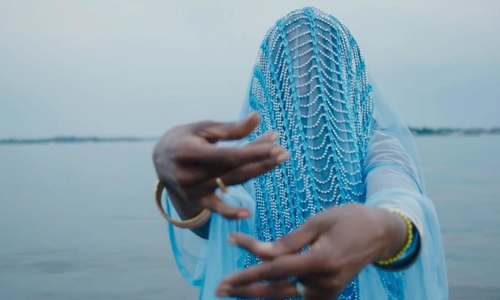 A photo still from the music video for Brujas by Princess Nokia. It shows a veiled woman standing in water, her arms outstretched towards the camera.