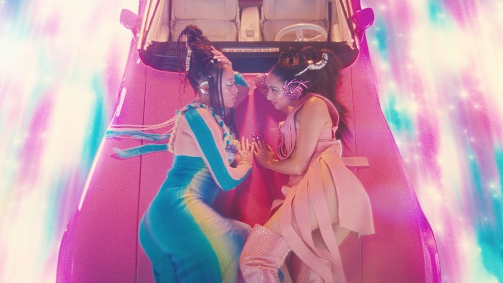 A photo still from the music video for Love Overgrown by Raveena. The image shows two women lying on the hood of a car looking lovingly into each other's eyes. The car is bright pink, and the rest of the colour palette is bright purple and blue.