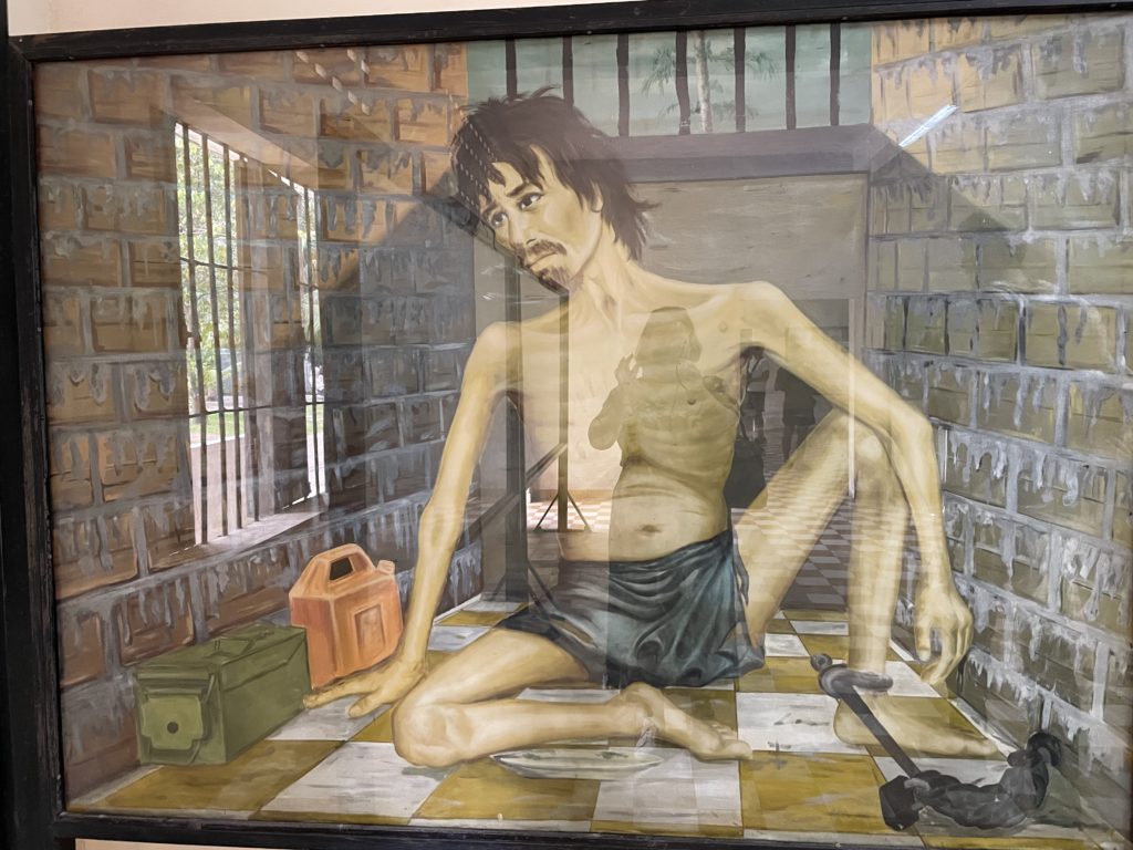 A photo of a painting by Vann Nath. The painting is of a starved prisoner sitting in a jail cell. His ribs are showing and his ankle is shackled to the wall.