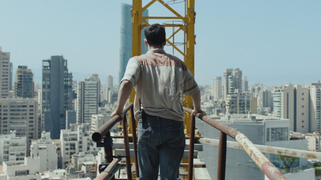 A photo still from the short film Warsha. The photo is of Mohammad walking across a metal bridge, his back to the camera, and the city of Beirut in the distance.