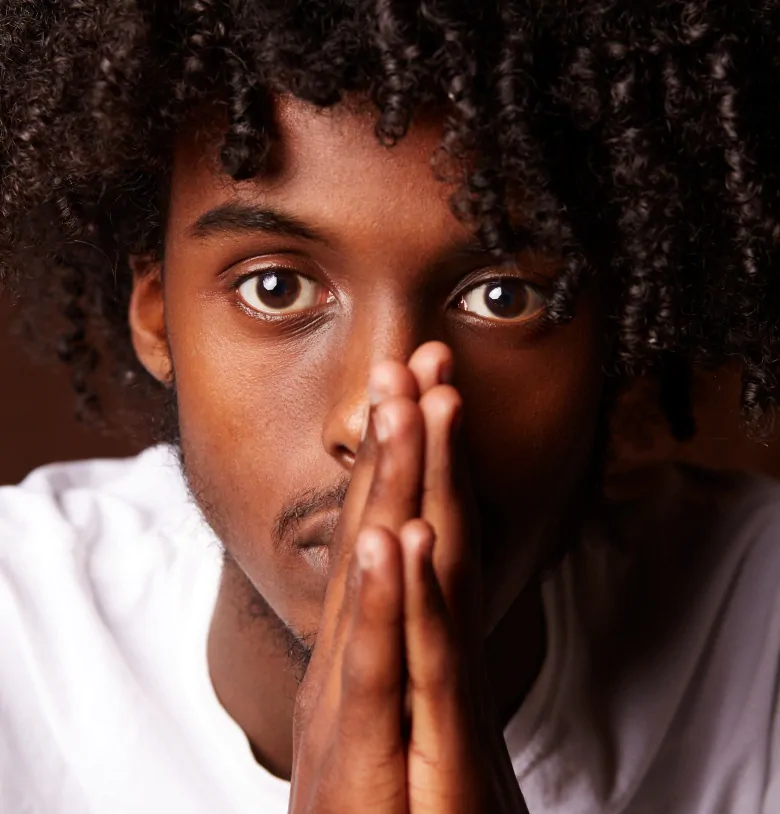 A photo from Faisa Omer's Portraits of Black Somali Youth from Ritchie. The photo is of a Black man looking straight into the camera with his hands pressed together in front of his face.