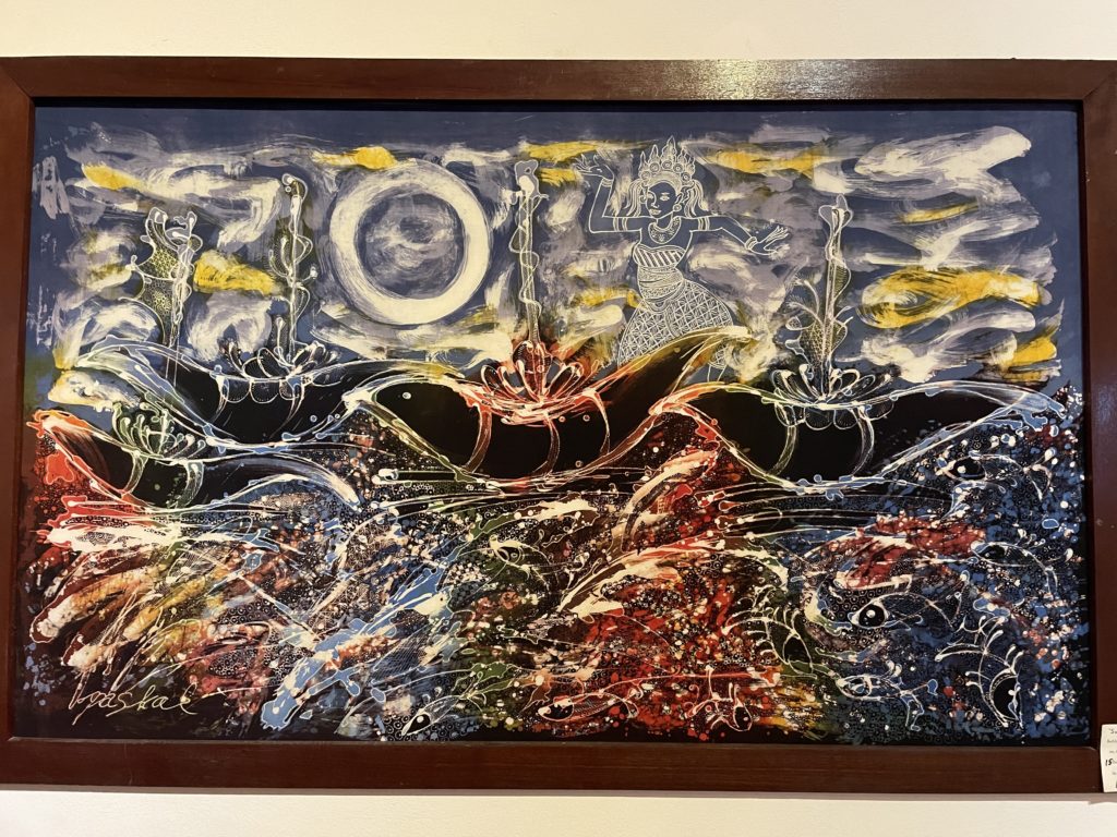A photo of a Batik canvas, on which an image of a goddess dancing on the waves of the ocean is depicted. The painting uses all the colours of the rainbow, but particularly blue and yellow.