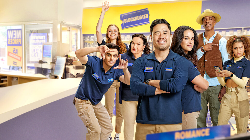 A promotional photo of the cast of Blockbuster. They are all in their Blockbuster t-shirts, adopting various character poses, looking straight into the camera.