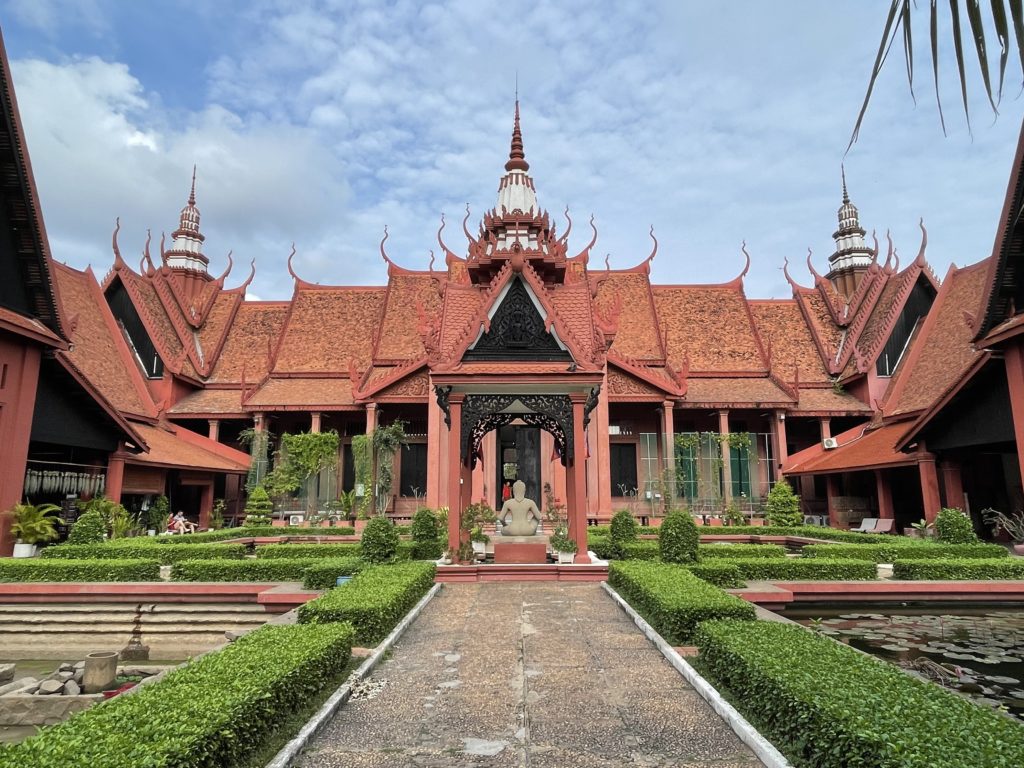 A photo of the exterior facade of the National Museum of Cambodia. There are buildings on either side, and a garden with a buddha statue in the centre.