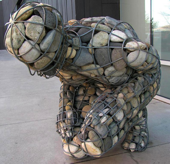 A photo of the artwork Rising Cairn by Celeste Roberge. The artwork is a metal outline of a human body, crouched down and hunched over, that is filled with stones.