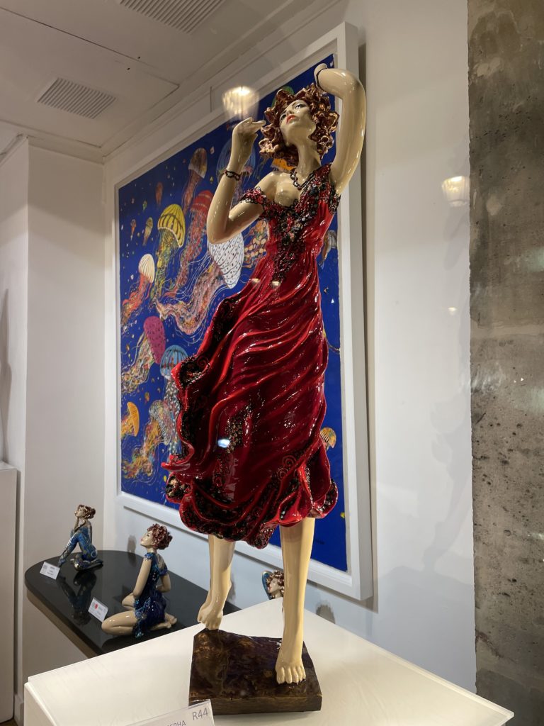 A photo of the statue Melia by the artist Josepha. It looks to be a porcelain figure of a woman wearing a red dress with her arms lifted towards the sky.