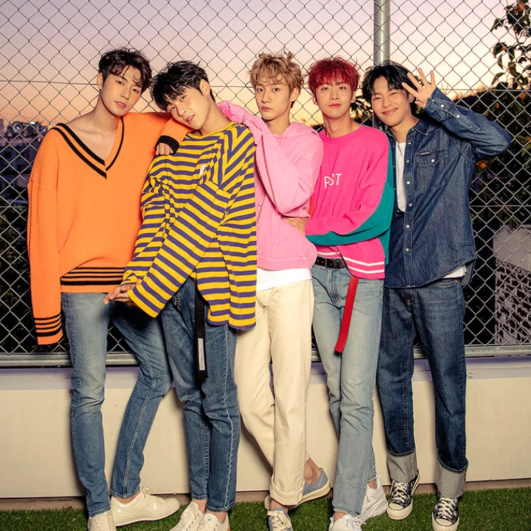 A group photo of the five members of KNK posing in front of a chain link fence and looking straight into the camera.