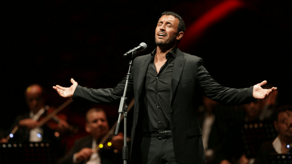 A photo of Kadim Al Sahir performing onstage during the Expo 2020 in Dubai. He is standing in front of a mic with his hands outstretched to his side.