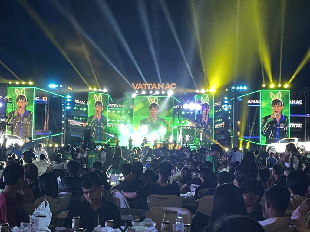 A photo of a concert stage in Kampot, Cambodia. A male singer wearing a black leather jacket and bunny ears is performing onstage.