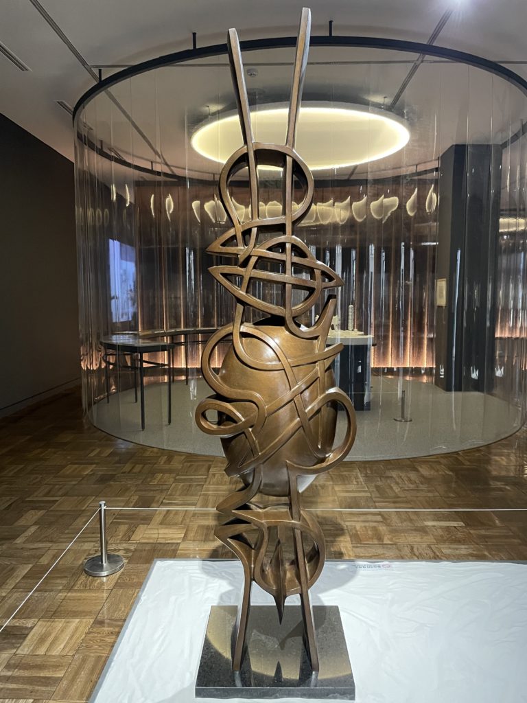 A photo of a statue by Moon Shin. The statue is made of bronze, is approximately two meters high, and consists of several abstract patterns of lines with a solid oval in the centre.