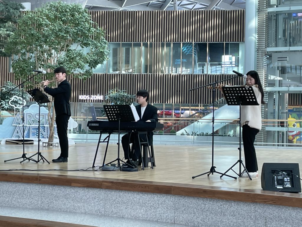 A photo of three musicians playing onstage at the Seoul airport. Two are playing the flute, while the third is playing a keyboard.