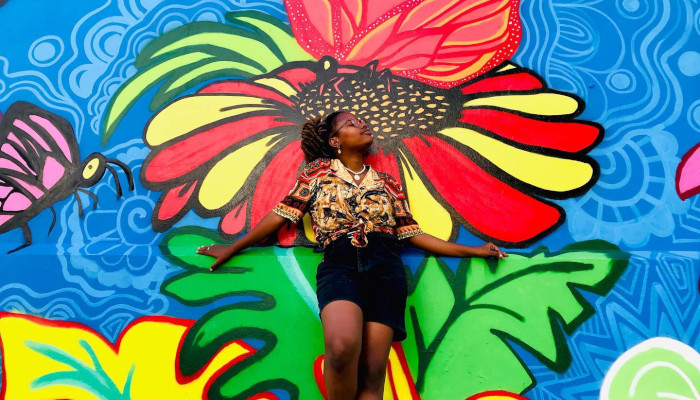 A photo of Vicky Onelien standing in front of one of her paintings. The painting is of a bright flower against a blue background.