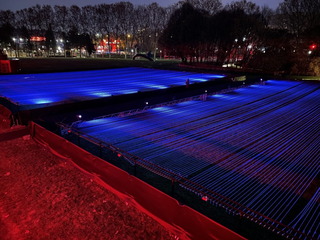A photo from above of the art installation by Yasuhiro Chida. It shows a field of illuminated parallel blue lines at night.