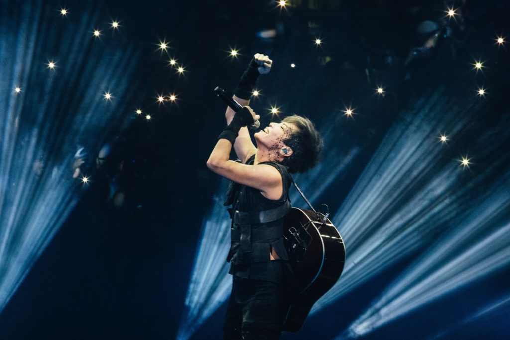 A photo of Denise Ho, onstage with blue lights behind her, a guitar slug on her back and a microphone in hand, singing with her face tilted to the sky.