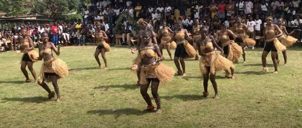 A photo still of the traditional dance from Gabon. Several dancers are dancing in a field wearing straw skirt costumes, surrounded by spectators.