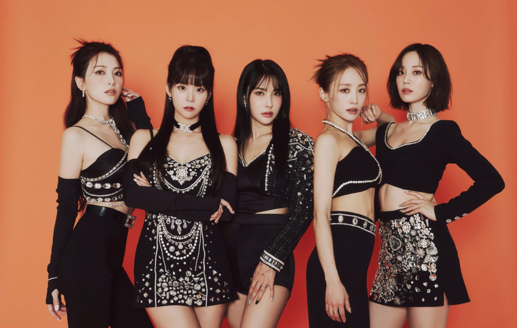 A promotional photo of the K-pop group Kara. The five members are standing in a line against an orange background, posing directly into the camera.