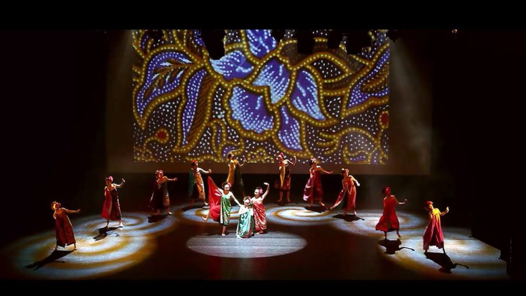 A photo still of the SMJK Phor Tay High School performing onstage. The photo is from a distance, showing the whole stage, including the 12 dancers mid-performance and the colourful backdrop behind them.