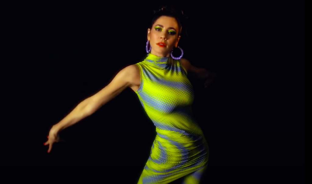 A photo still of Marina from the music video of Purge Your Poison. She's wearing a patterned, lime green dress and posing straight into the camera against a black background.