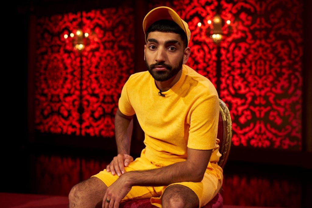 A photo of Mawaan Rizwan sitting on a bench with a red background, staring straight into the camera.