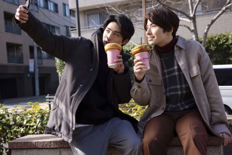 A photo still from Old Fashion Cupcake. Nozue and Togawa are sitting outside, taking a selfie while holding donut drinks.