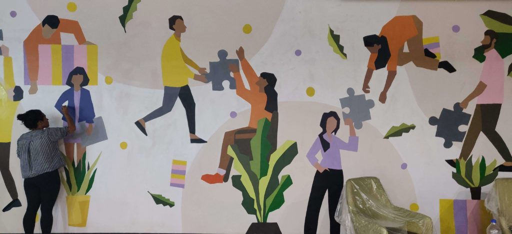 A photo of a mural by the Aravani Art Project. It shows various people with abstract faces handing each other large puzzle pieces.