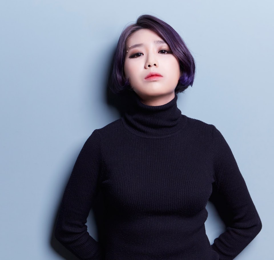A promotional photo of Yeeun Ahn. She is looking straight at the camera, leaning up against a pale blue wall.