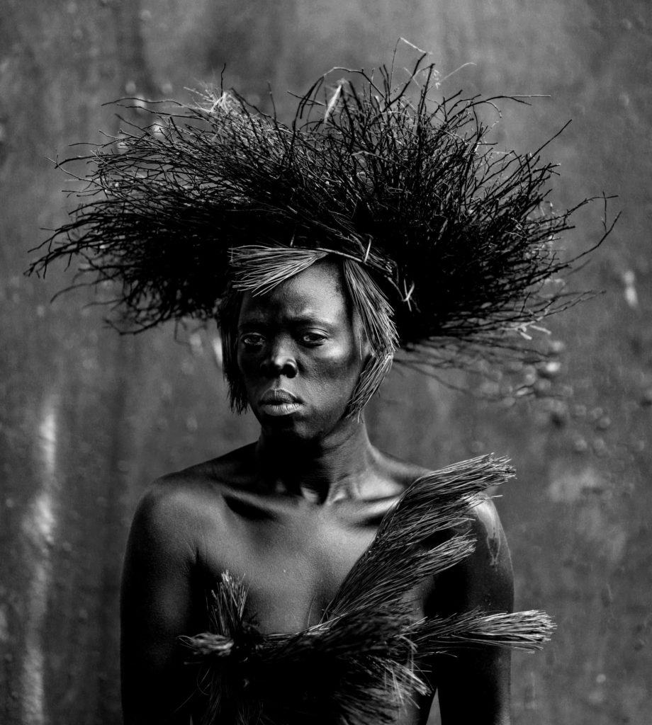 A black and white self portrait of Zanele Muholi. They are wearing a dress and hat made of straw.