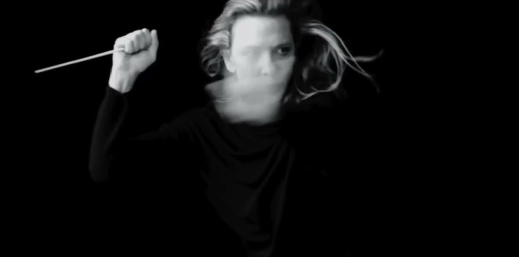 An image showing a woman holding a conductor's wand. Her face is slightly blurred as if she's shaking her head too fast.
