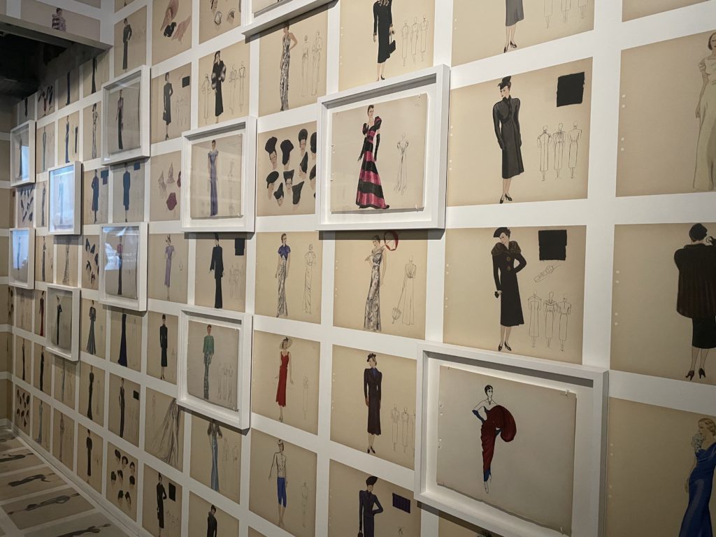A photo showing a wall of mounting and framed drawings by Elsa Schiaparelli. Each drawing is of a different dress.