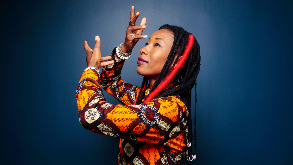 A photo of Fatoumata Diawara. She is posing in front of a blue wall in a colourful outfit. She is in profile with her hands held up in front of her face.