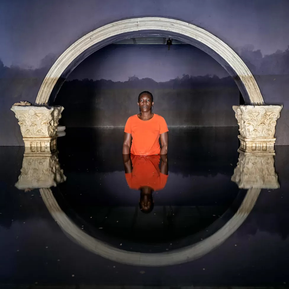 A photo by Gideon Mendel. It shows a man standing in a doorway to a building, submerged up to his waist in water.