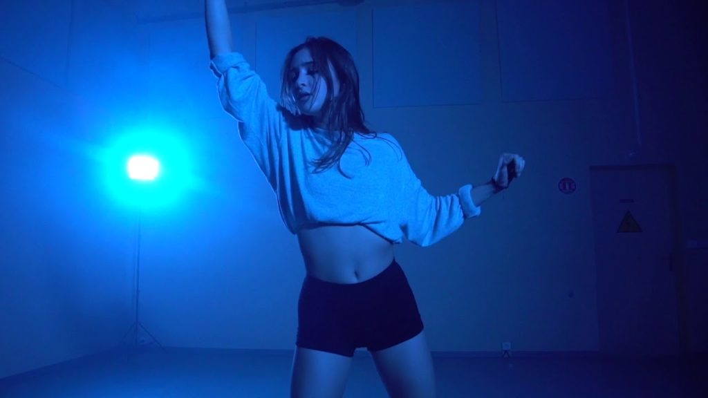 A photo still of Loriane Cateloy-Rose, mid performance. She is wearing a white top and black shorts. One arm is outstretched above her and she is bathed in blue light.