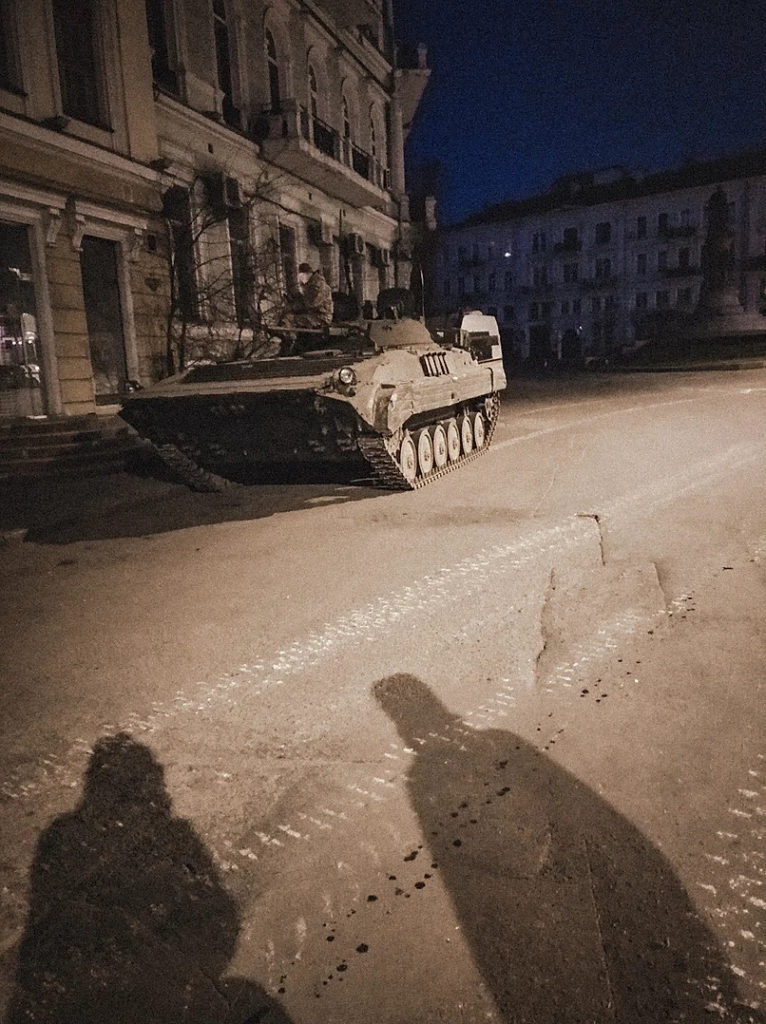 A photo by Maryna Semenkova. It shows a city street in Ukraine at night, with a Russian tank sitting across the road.