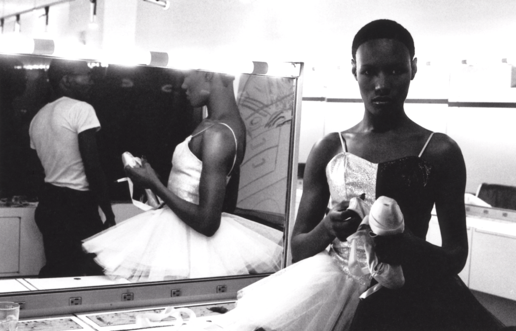 A photo by Ming Smith. It shows a Black ballerina in a tutu and holding ballet shoes. She is standing in front of a mirror and staring straight into the camera.