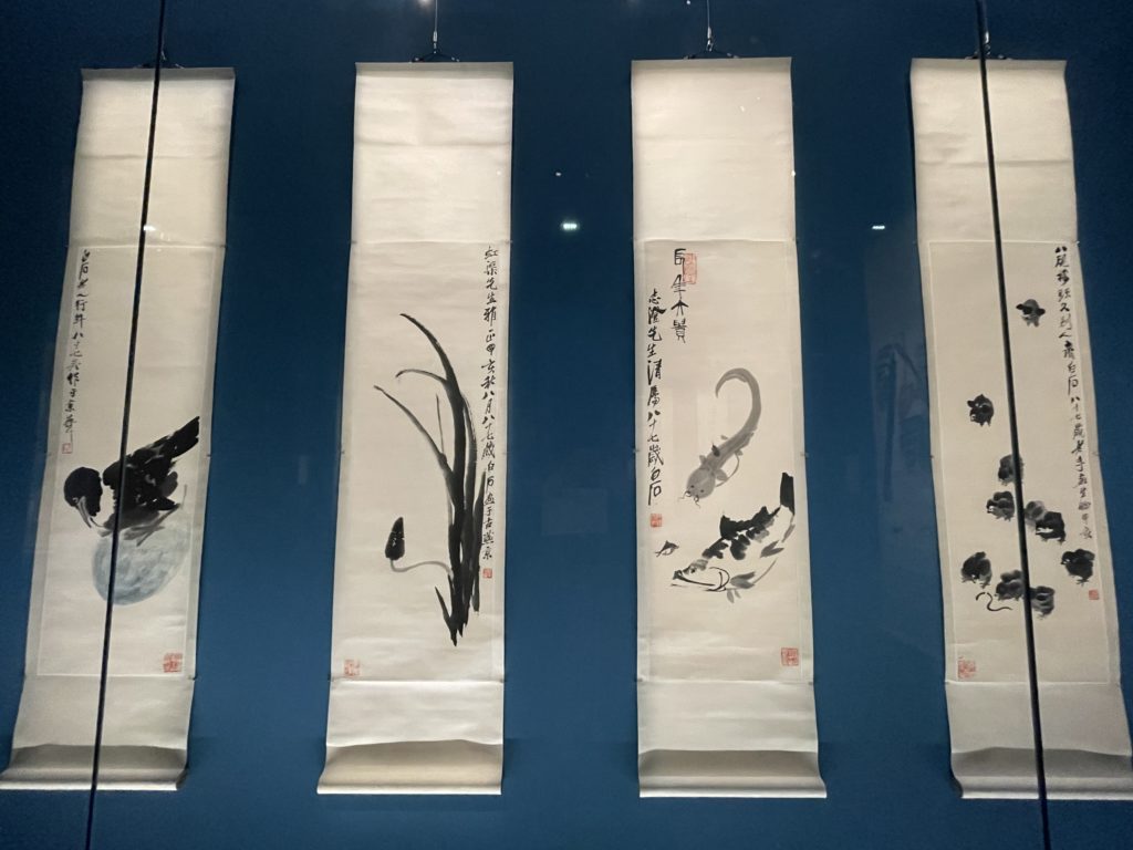 A photo of four vertical ink paintings by Qi Baishi. One is of a bird, one is of a reed-like plant, one is of two fish, and one is of a group of chicks.