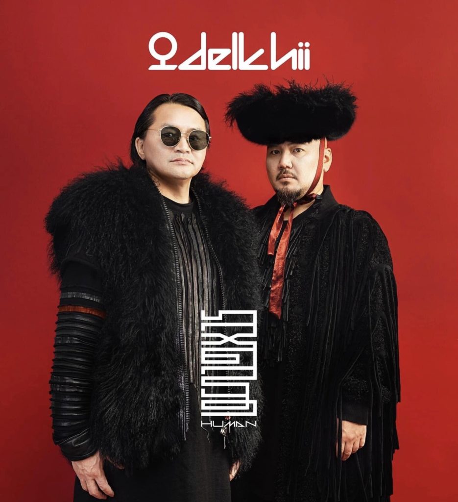 A photo of the two lead singers of The Delkhii band. They are standing side by side against a red backdrop, staring straight into the camera.