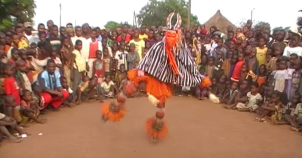 A photo of a Zaouli de Manfla dancer mid-performance. He is wearing an orange mask and is surrounded by a circle of villagers.