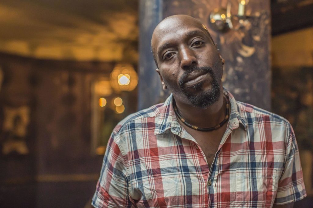 A photo of Kei Miller from the chest up, staring straight into the camera, head slightly tilted to the left.