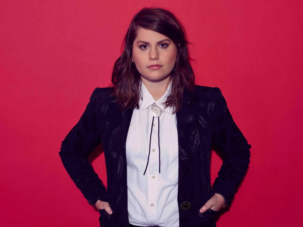 A photo of Alex Lahey staring straight into the camera, posed against a red wall.