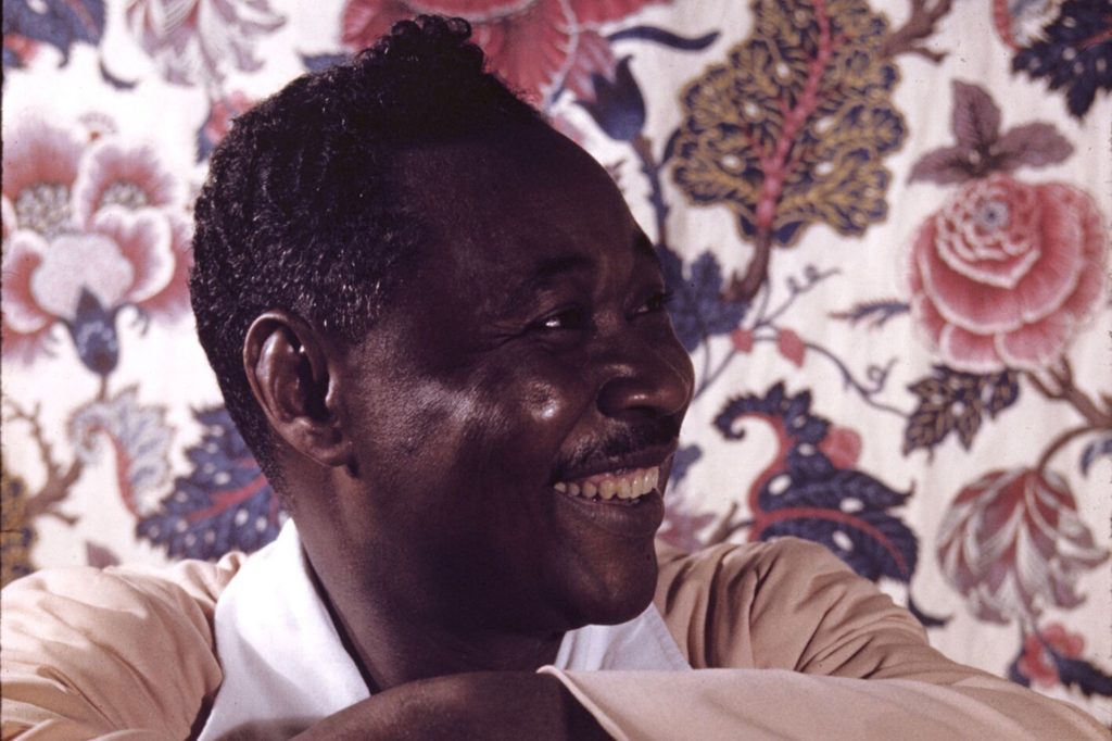 A profile photo of Claude McKay. He is sitting in front of a floral background and smiling while looking off camera to the right.