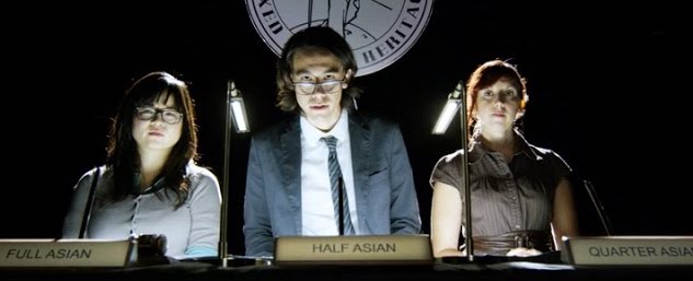 A photo still from the sketch Are You Asian Enough? It shows three people behind a raised desk, staring down at the camera.