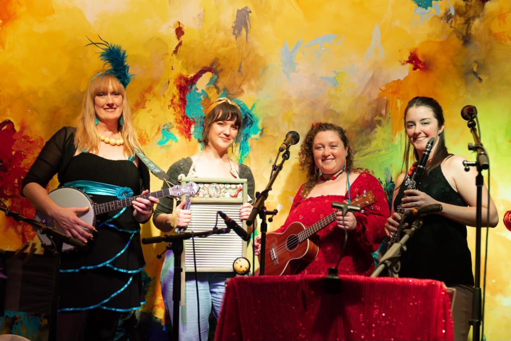 A promotional photo of the Hot Mamas. The four women are standing in a line, holding their instruments, and smiling straight into the camera.