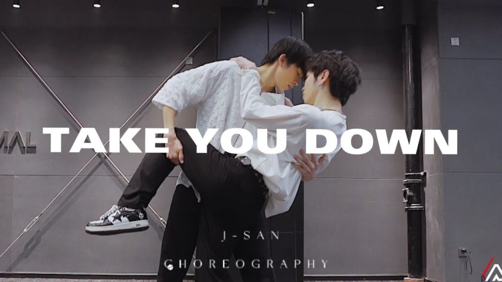 A photo still from a video by choreographer J-San. It shows two male dancers, mid-performance, one holding the other in his arms.
