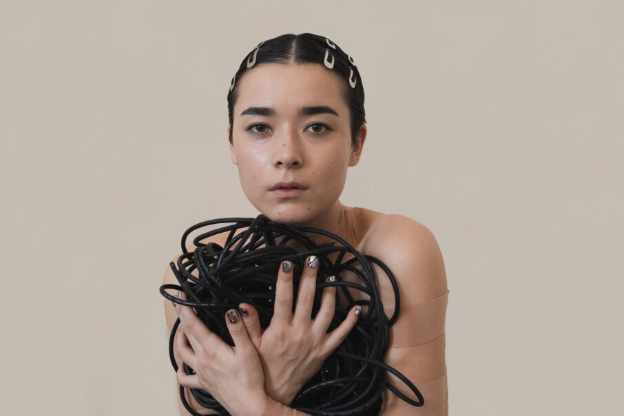 A photo of singer Miss Grit. She is posed in front of a beige background, looking straight into the camera, holding a bundle of rope to her chest.