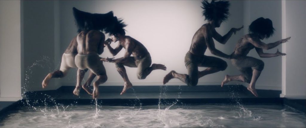 A photo still from the music video for "Oui mais... Non" by Mylène Farmer. It shows several dancers mid-leap in the air above a pool of water.