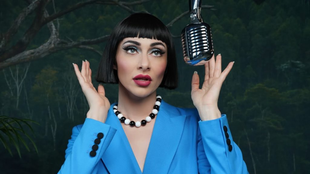 A photo still from the video for Chucky Cheese by Qveen Herby. She is standing in front of a recording mic, wearing a bright blue top, hands up framing her face.