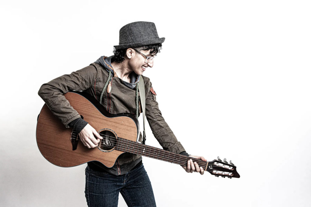 A photo of Shireen Amini. They are standing in profile against a white background holding a guitar.