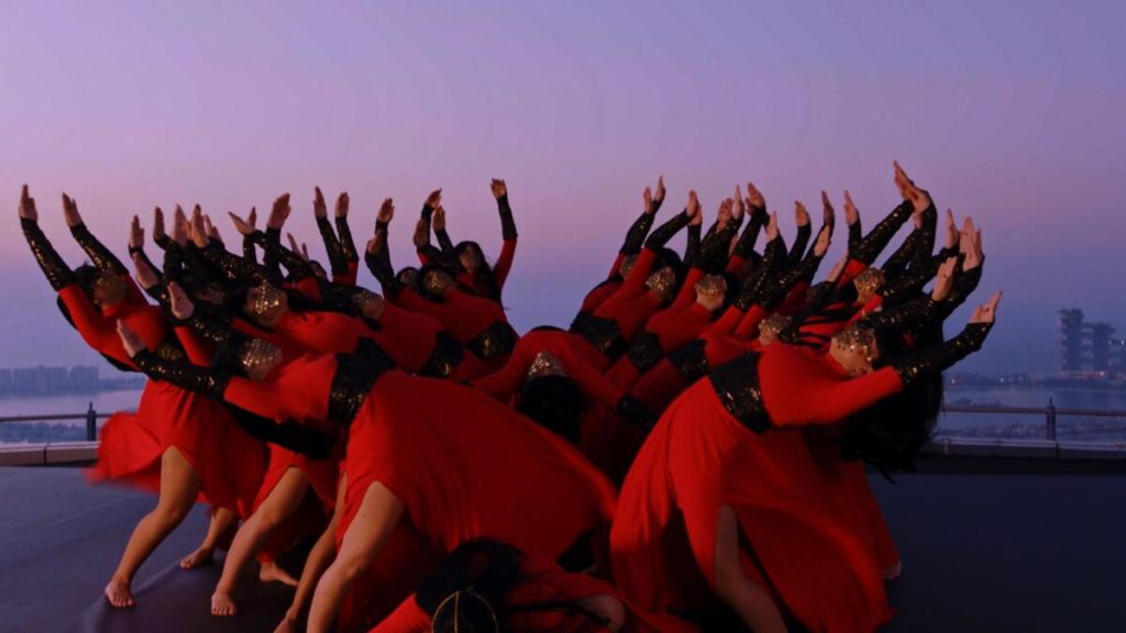 A photo still from a performance of the Mayyas. A group of the dancers are outside, dressed in red, with their arms raised in the air.