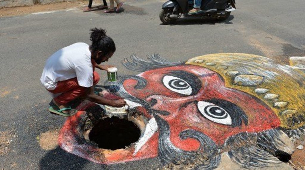 A photo of Baadal Nanjundaswamy. He is crouched over one of his street art pieces, painting the image. The image is of a stylized figure where the mouth is a pothole in the road.