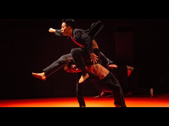 A photo still from the piece Judo by the Bereishit Dance Company. Two dancers are onstage. One is jumping over the other.